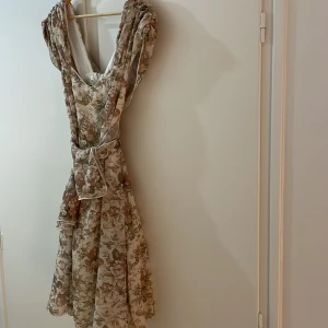 Finklänning - Blommig klänning med tyll i kjoldelen, dragkedja i rygg och 2 sleeves från axlarna som kan knytas runt midjan, hals eller hänga fritt.  Tyget har lossnat inuti vid dragkedjan, men det varken syns eller känns