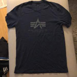 En mörkblå t-shirt från Alpha Industries med deras logotyp tryckt på framsidan. T-shirten har korta ärmar och en rund halsringning.