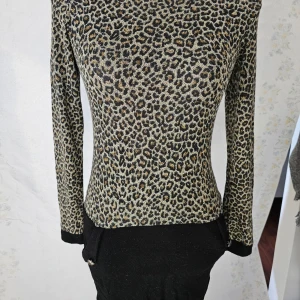 Leopardmönstrad tröja från Mega Collector - En leopardmönstrad klänning. Klänningen har långa ärmar och en stretchig passform. Den är huvudsakligen i leopardmönster med svarta detaljer vid ärmslut och nederkant.