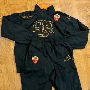 Roma Kappa Vintage 00’s Set Jacka och Byxor - Sjukt coolt set från tidigt 00-tal när Roma hade Kappa som leverantör. Tyvärr har de inte Kappa längre men här finns iallafall chansen att plocka hem ett galet snyggt set i perfekt skick med jacka och långa byxor.   Kolla in mina andra annonser.