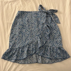 kjol - ljusblå kjol med vita blommor, har volang i nederkanten, samt en likande detalj som följer upp till knytningen, dragkedja bak. inga defekter och perfekt till sommaren🌸