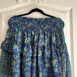 Blå mönstrad kjol med volanger - Säljer en superfin blå kjol med ett detaljerat mönster i grönt och beige. Kjolen har volanger som ger den en extra touch och en elastisk midja för en bekväm passform. Perfekt för sommarens alla fester eller en dag på stan!