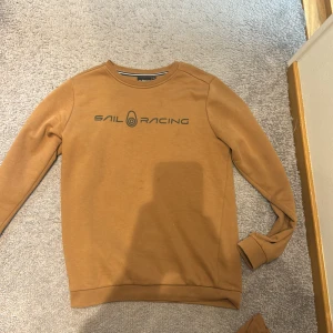 Brun tröja från Sail Racing - Säljer en snygg brun tröja från Sail Racing i storlek 170. Tröjan är gjord av bomull och har en normal passform. Den har långa ärmar och ett tryck med Sail Racing-loggan på framsidan. Perfekt för höst och vinter!