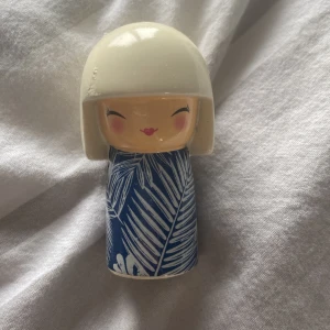 Söt docka från Skincity - En söt liten docka från Skincity med en vit bobfrisyr och ett blått klänning med vita bladmönster. Perfekt som en liten dekoration eller present. Dockan har ett glatt ansikte med rosiga kinder och röda läppar. Den är tillverkad med kärlek och är miljövänlig.