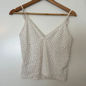 Vit blommig linnetopp från Brandy Melville - En söt och somrig vit linnetopp från Brandy Melville med små blommor i rosa och grönt. Toppen har smala justebara axelband och en v-ringad halslinje. Perfekt för varma sommardagar.