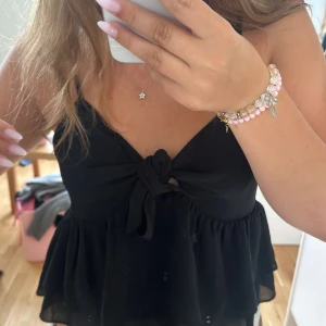 Svart topp med knytning - En svart topp  med knytning framtill och smala axelband. Toppen har en volangdetalj vid midjan. Passar både till vardag och fest🫶🏻💘💞🌅