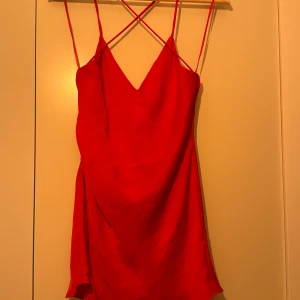 Röd kortklänning med smala axelband - Säljer en superfin röd kortklänning med smala axelband. Klänningen har en v-ringad fram och en snygg öppen rygg med korsade band. Den är perfekt för fest och sommar, och materialet känns som satin vilket ger en lyxig känsla. Klänningen har en liten slits på sidan som gör den extra chic. Passar perfekt för en utekväll eller en speciell tillställning!