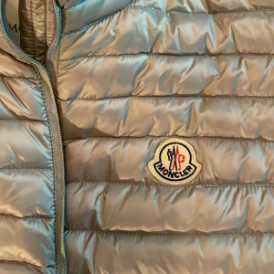 Äkta Moncler Väst Dupre Turtleneck grå/beigh/silver aktig. - En beige/grå aktig Moncler väst med lite glans i tyget, modellen är Dupres Turtleneck. Inga defekter någonstans då den bara varit i garderoben sen den köptes! Som ny. Finns streckkod i som man kan kolla på hemsidan så den är äkta. Perfekt till våren!