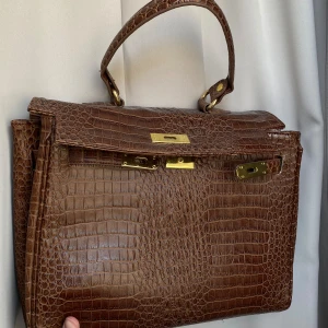Brun väska i Kelly style - Jätte fin brun Kelly inspirerande väska, den enda defekten är att själva pinnen for att kunna stänga väskan har ramlat lös (se sista bilden) men väskan går fortfarande att andvända och om man vill kan man sätta dit den igen. Axelband ingår🤎