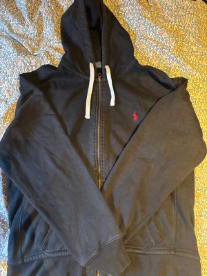 Svart hoodie från Polo Ralph Lauren - Svart hoodie från Polo Ralph Lauren med vit snörning i huvan och röd broderad logga på bröstet. Tröjan har dragkedja framtill, långa ärmar och två fickor. , perfekt för chill dagar och streetwear.