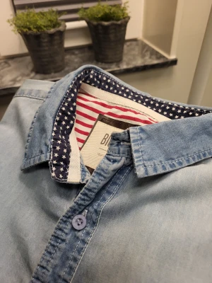 Ljusblå jeansskjorta från Big Star - Ljusblå jeansskjorta från Big Star med lång ärm, bröstficka och coola detaljer som stjärnmönstrat tyg på insidan av kragen och rödvita ränder. Skjortan har knappar framtill och diskreta rutiga detaljer på ärmarna.