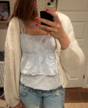 Ljusblå volangig linnetopp - Supersöt ljusblå linnetopp med volanger och smock upptill. Toppen har tunna axelband med spetsdetalj och ett skirt, lätt glansigt tyg. Perfekt för varma dagar och ger en romantisk vibe till outfiten.