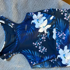 Blommig linnetopp från Stronger - Supersnygg linnetopp från Stronger med blommigt mönster i blått, vitt och rosa. Toppen har en sportig racerback-design och är tillverkad i mjuk polyester som andas. Perfekt för träning och ger en fräsch vibe med sina tropiska blommor och blad.
