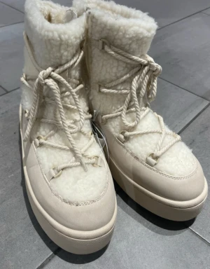 Vita vinterskor - Säljer ett par helt nya snygga beige moon boots med grov snörning och platt sula. Skorna har en fluffig, pälsliknande ovandel och detaljer i skinn som ger en cool vinterlook. Perfekta för dig som vill ha både stil och värme under kalla dagar.