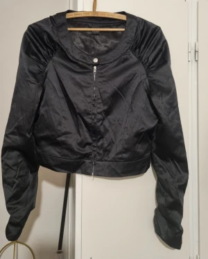 Svart croppad jacka med dragkedja - Snygg svart croppad jacka från Vila med rund halsringning och långa ärmar. Jackan har en diskret dragkedja framtill och är tillverkad i ett glansigt syntetmaterial som ger en cool look. Perfekt för dig som gillar stilrena och trendiga plagg.