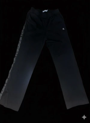 Svarta byxor från Calvin Klein Jeans - Svarta byxor från Calvin Klein Jeans med elastisk midja och raka ben. Snygg logotyp längs sidorna och liten CK-brodyr framtill. Materialet är mjukt och har en sportig känsla, perfekt för chill eller streetwear. Passformen är normal och stilen är straight.