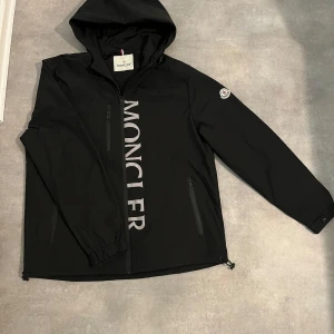 Svart vindjacka från Moncler - Svart vindjacka från Moncler med stor logotyp framtill och klassisk patch på ärmen. Jackan har huva, två fickor med dragkedja och justerbar nederkant. Materialet är lätt och syntetiskt, perfekt för blåsiga dagar. Snygg och stilren design med lång ärm och hel dragkedja.