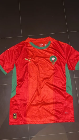 Marocko fotbollströja Puma röd/grön - Säljer en officiell Marocko fotbollströja från Puma i rött med gröna detaljer och guldigt Puma-logga. Tröjan har korta ärmar, rund hals och broderat Marocko-emblem på bröstet. Tillverkad i lätt polyester med snyggt mönster i tyget.