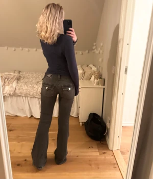 Grå/svarta bootcut jeans från True Religion - Säljer dessa superpopulära jeans från True Religion i modellen Joey Low Rise Flare Flap i färgen Charcoal. Dessa är verkligen mina drömjeans så tar emot att sälja, men de ligger bara helt oanvända i garderoben och samlar damm eftersom de är lite för långa på mig. De är alltså helt oanvända och endast provade.