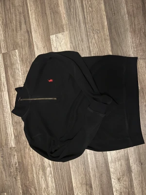 Svart half zip tröja från Polo Ralph Lauren - Svart långärmad tröja från Polo Ralph Lauren med half zip och klassisk röd logobrodering på bröstet. Tröjan har en hög krage och är gjord i mjuk bomull, perfekt för chill dagar. Snygg och enkel att matcha med jeans eller joggers.
