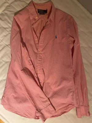 Rutig rosa skjorta Polo Ralph Lauren - Slim fit skjorta från Polo Ralph Lauren i storlek S. Skjortan är rosa med vitt rutmönster och har klassisk krage, knappar framtill och lång ärm. Broderad blå Polo-logga på bröstet. Tillverkad i mjuk bomull, perfekt för dig som gillar stilrena och fräscha plagg.