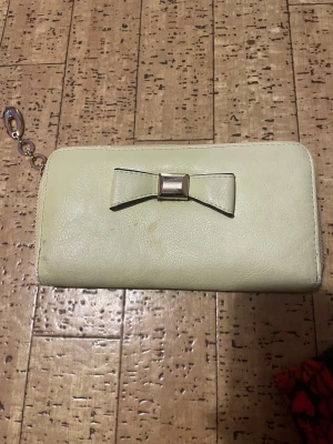 Ljusbeige plånbok med rosett - Snygg ljusbeige plånbok med dragkedja och dekorativ rosett i metall på framsidan. Plånboken har flera fack för kort och mynt, samt en kedja med metallring för att fästa nycklar eller accessoarer. Perfekt storlek för att ha med sig överallt.