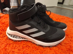 Svarta Adidas sneakers med kardborre - Svarta Adidas sneakers med reflekterande detaljer och vita ränder på sidorna. Skorna har både snörning och kardborreband upptill för extra support. Grov sula med bra grepp, perfekt för höst och vinter. Tillverkade i syntetmaterial och textil.