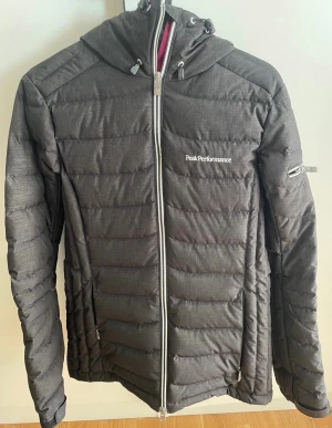 Mörkgrå Peak Performance dunjacka S - Peak Performance dunjacka i mörkgrått med huva och vita detaljer. Jackan har quiltad design, dragkedja framtill och på ärmen, samt logga på bröstet och ryggen. Perfekt för kalla dagar och snygg till streetwear. Fodret har färgglada band i blått, gult och rosa.