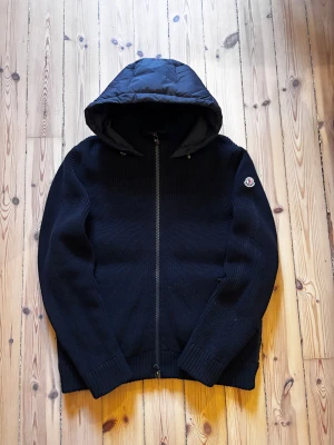 Mörkblå Moncler cardigan - Mörkblå cardigan från Moncler med stickade ärmar och framstycke, samt quiltad rygg och huva. Jackan har fickor med dragkedja. Perfekt för dig som vill ha en snygg och varm cardigan. Storlek L sitter som M. Skick 9/10. Fråga på!