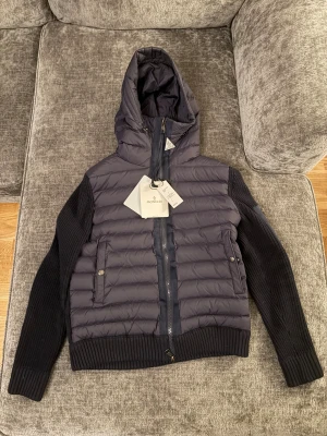 Svart Moncler pufferjacka med huva - Snygg marinblå pufferjacka från Moncler med stickade ärmar och quiltad kropp. Jackan har huva, dragkedja framtill och två fickor med tryckknappar. Materialmix av dun, polyester och stickat ger en modern vibe. Triangel taggen ligger ner på jackan då snörret är sönder.