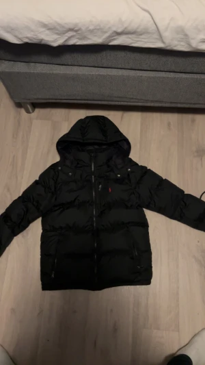 Svart pufferjacka från Polo Ralph Lauren - Svart pufferjacka från Polo Ralph Lauren med huva och dragkedja framtill. Jackan har två sidofickor med dragkedja och en liten röd logga på bröstet. Perfekt för kyliga dagar och riktigt skön att bära.