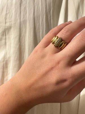 Guldring med glittriga stenar - Snygg guldfärgad ring med tre breda band och ett mittparti täckt av glittrande stenar. Ringen har en modern och cool design som sticker ut på handen. Perfekt för dig som gillar att addera lite extra bling till din stil.