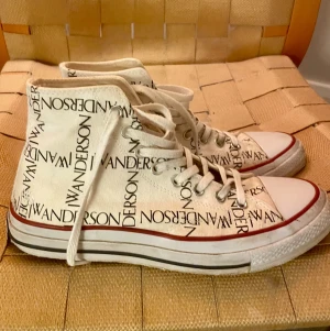 JW Anderson x Converse vita high-top - Unika vita high-top sneakers från Converse i samarbete med JW Anderson. Skorna har svart logotyp-tryck över hela canvasen, klassisk rund tå, snörning och tjock vit sula med röd rand. Perfekta för dig som vill sticka ut med en cool och trendig look.