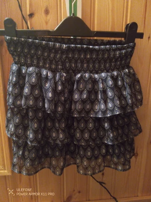 Mönstrad volangkjol från Lindex - Snygg kortkjol från Lindex med mörkblå botten och droppformade mönster i blått och vit. Kjolen har flera lager volanger och en bred, smockad midja som ger en skön passform. 