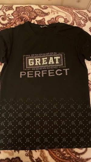 Svart t-shirt med stenar från Caceek - Svart t-shirt från Caceek med texten 'GREAT PERFECT' framtill i glittriga stenar och dekorativa mönster. Nedre delen har prickigt mönster och tryckta bokstäver. Klassisk rund hals och korta ärmar. Perfekt för dig som gillar statement-plagg.