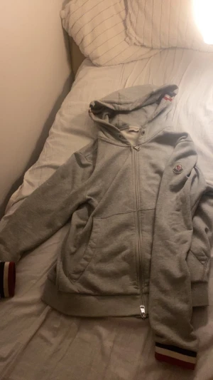 Grå hoodie från Moncler med dragkedja - Säljer en grå hoodie från Moncler med hel dragkedja och huva. Tröjan har Moncler-logga på bröstet och ribbade muddar med röd, vit och blå rand. Perfekt för chill dagar och snygg streetstil. Storlek m men passar S som perfekt