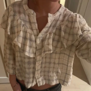 Rutig beige skjorta med volang - Säljer en beige rutig skjorta med volangdetaljer över axlar och bröst. Skjortan har knappar framtill och är croppad med trekvartsärm. Materialet känns mjukt och luftigt, perfekt för dig som gillar en chill och trendig look.