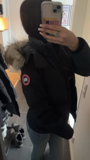 Svart dunjacka från Canada Goose - Säljer en svart dunjacka från Canada Goose med klassisk patch på ärmen och stor huva med fluffig fuskpäls. Jackan har en rak passform och är perfekt för kalla vinterdagar. Snygg och tidlös design som håller dig varm hela säsongen.