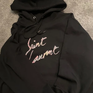 Svart hoodie  - Säljer en stilren svart hoodie från Saint Laurent med deras ikoniska logga i en färgglad design på bröstet. Perfekt för en avslappnad och trendig look. Hoodien har en klassisk passform med dragsko i huvan. Passar XS