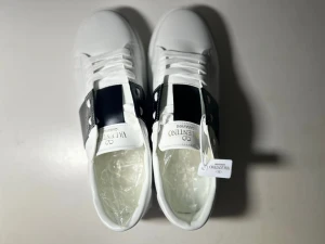 Valentino Garavani vita sneakers - Snygga vita sneakers från Valentino Garavani med svart detalj på sidan och klassisk logga på plösen. Skorna har snörning, platt sula med mönstrad undersida och är tillverkade i skinn för en lyxig känsla. Perfekt för dig som gillar stilrena och exklusiva sneakers.