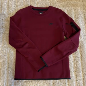 Nike Tech Fleece sweatshirt  - Snygg vinröd långärmad tröja från Nike med svart logga på bröstet och en diskret ficka med dragkedja på ärmen. Tröjan har rund hals och svarta detaljer vid muddar och nederkant. Materialet är mjukt med 66% bomull och 34% polyester.