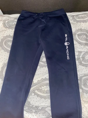 Mörkblå mjukisbyxor Sail Racing - Mörkblå mjukisbyxor från Sail Racing med vit logga längs benet. Byxorna har snörning i midjan, fickor på sidorna och en bakficka. Sköna och stilrena, perfekta för chill eller träning. Materialet är mjukt och känns som bomull.