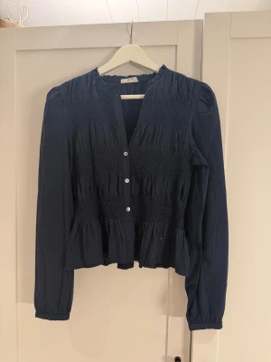 Mörkblå blus med smock och knappar - Supersnygg mörkblå blus från mango med smockade partier och volang nedtill. Blusen har rund hals, långa ärmar och knappar framtill i kontrasterande färg. Perfekt för dig som gillar en feminin och trendig look. Storlek barn:M men motsvarar S