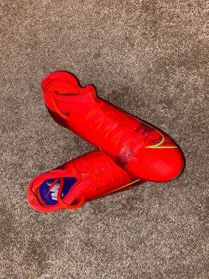 Nike röda fotbollsskor med snörning - Säljer ett par grymma Nike fotbollsskor i starkt röd färg med coola gröna och svarta detaljer. Skorna har snörning och en platt sula med dobbar i orange och silver, perfekt för spel på gräs. Materialet är syntet och designen är riktigt sportig och modern.