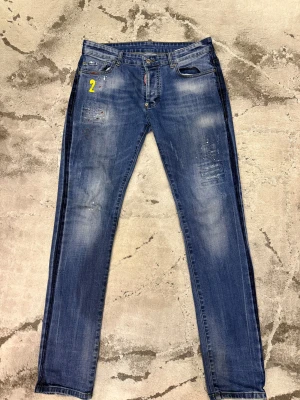 Dsquared2 Jeans - Snygga blå jeans med slitningar, färgstänk och ett gult tryck med siffran 2 framtill. På bakfickan finns broderade detaljer med flagga, glas och burk. Jeansen har en rak passform och klassisk femficksdesign. Perfekt för dig som gillar unika detaljer och streetstyle.