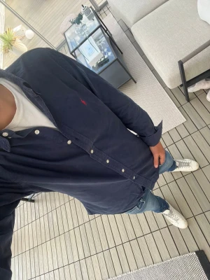 Mörkblå skjorta Polo Ralph Lauren - Snygg mörkblå skjorta från Polo Ralph Lauren i slim fit. Skjortan har klassisk krage, vita knappar och den ikoniska röda logon broderad på bröstet. Tillverkad i mjuk bomull, perfekt för dig som gillar stilrena och tidlösa plagg.