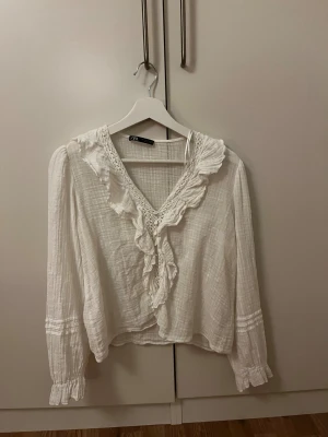 Vit blus med volang från Zara - Supersöt vit blus från Zara med v-ringning, volangdetaljer och spets längs halsringningen. Långärmad modell i lätt, luftig bomull med rynkade ärmslut och romantisk känsla. Perfekt för dig som gillar en feminin och trendig stil.