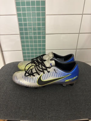 Nike Mercurial silver/blå fotbollsskor neymar - Nike Mercurial fotbollsskor i silver med blå och gula detaljer, svarta snören och svarta dobbar. Används och gammal modell men inte några skador mer en smutts vilket enkelt går bort. Hör av dig vid frågor ( priset kan diskuteras)