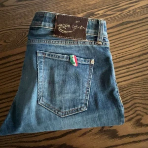 Blå jeans från Jacob Cohen - Snygga blå jeans från Jacob Cohen med klassisk femficksdesign och en liten italiensk flaggdetalj på bakfickan. Jeansen har en normal passform och är tillverkade i mjukt denimtyg med lätt slitning för en cool look. Dem sitter otroligt bra men är för stora så därför är dem till salu. Hör av dig vid frågor🏆