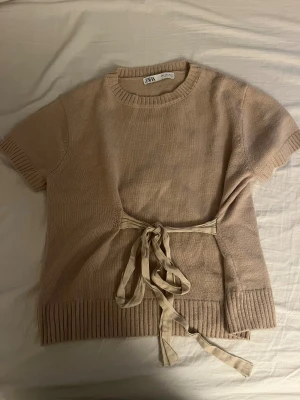 Beige stickad tröja med knyt från Zara - Säljer en trendig beige stickad tröja från Zara med korta ärmar och rund hals. Tröjan har ett snyggt knytband i midjan som ger en cool detalj och ribbade muddar vid ärmslut och nederkant. Säljer den för att den inte får till användning mer. Tröjan är liten i storlek o skulle säga att den passar storlek S!💕💕